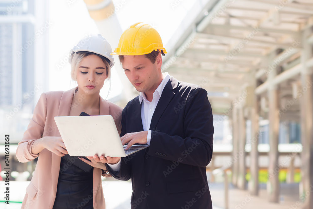 Male and female engineers wearing protective caps are looking at building construction plans from a notebook in the outdoors. With a backdrop to the building structure Male and female engineers wearing protective caps are looking at building construction plans from a notebook in the outdoors. With a backdrop to the building structure