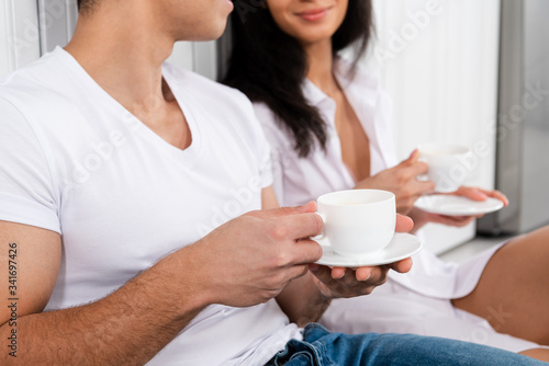 Cropped view of interracial couple with saucers and cups of coffee in kitchen