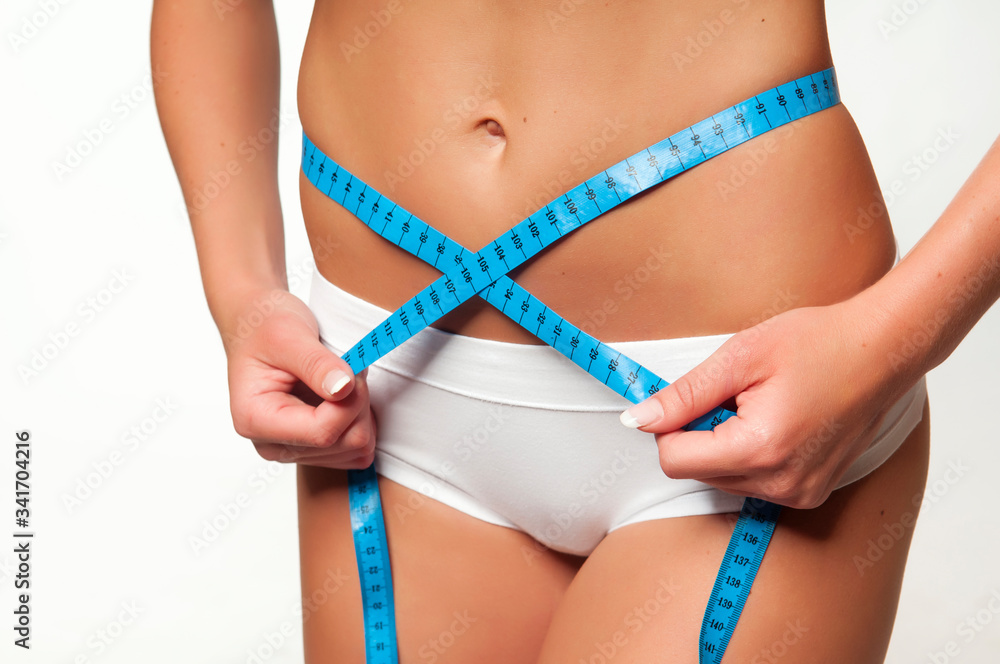 Weight loss - woman with trained belly. Young skinny female in white panties with tanned body is measuring her waist with blue tape measure and showing diet results. Studio shot, white background