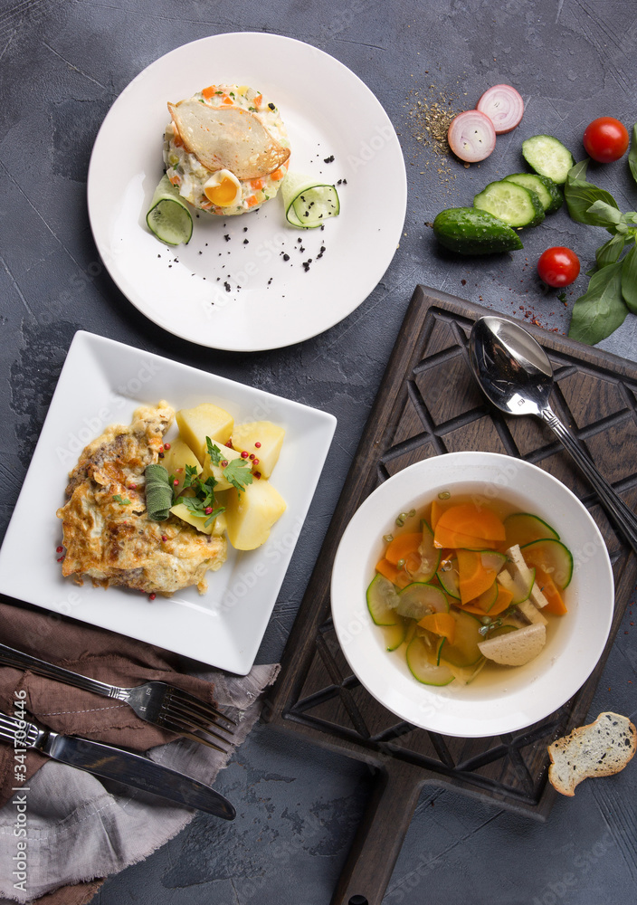 Complex dinner. Vegetable soup, salad Olivier and potatoes with fish on white plates on a dark gray background. Fresh vegetables and appliances. Top view, flatlay. Background image, copy space Complex dinner. Vegetable soup, salad Olivier and potatoes with fish on white plates on a dark gray background. Fresh vegetables and appliances. Top view, flatlay. Background image, copy space