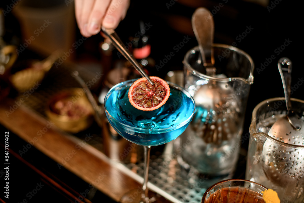 close-up of glass with blue cocktail that bartender's hand decorates with slice of citrus
