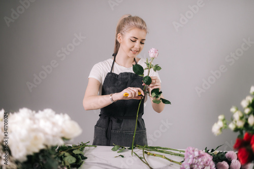 Beautiful young woman florist wearing apron working with fresh rose at the table on white background. Concept of working with flowers, floral business.