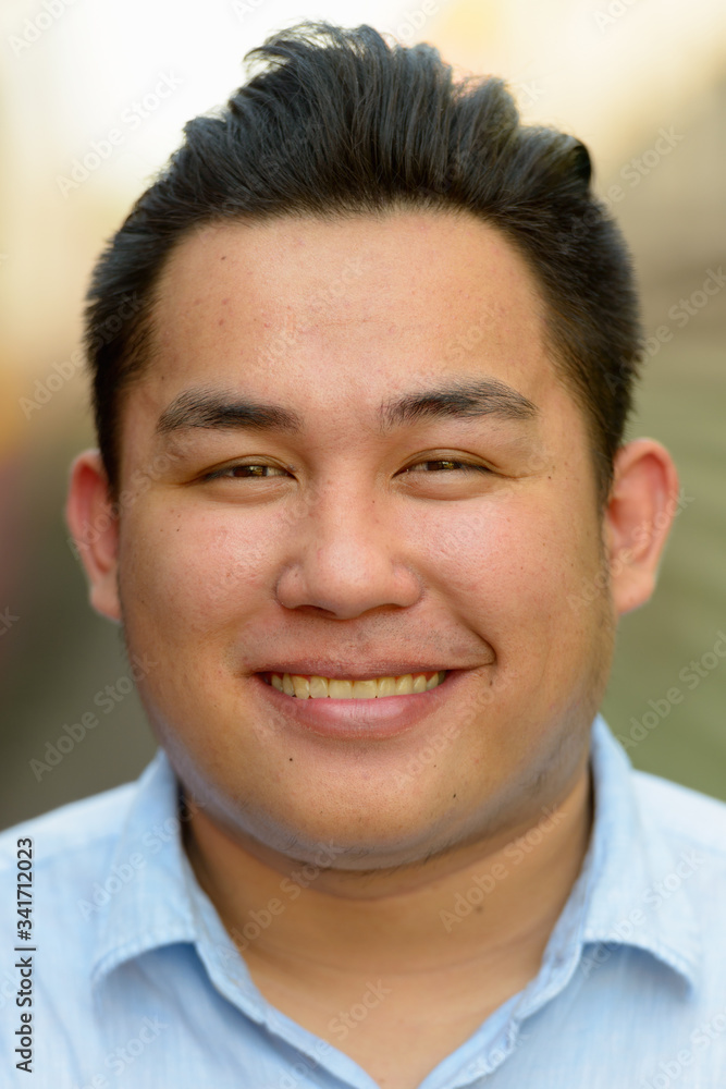 Face of happy overweight Asian tourist man smiling at railway train station Face of happy overweight Asian tourist man smiling at railway train station