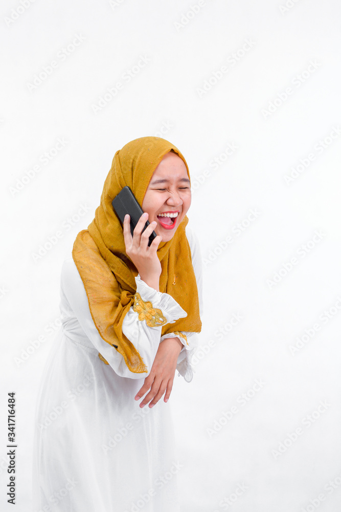 portrait of attractive asian muslim woman on white dress with hijab using a smartphone and laugh while have a call isolated over white background portrait of attractive asian muslim woman on white dress with hijab using a smartphone and laugh while have a call isolated over white background