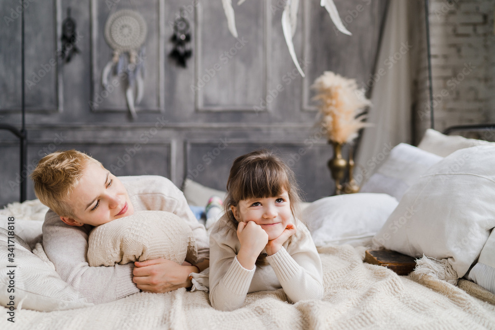 Mother and daughter hugging and cuddling in bed. Pretty little girl and beautiful woman with short haircut lie together in stylish bedroom. Family weekend, beauty day, having fun, love concept.