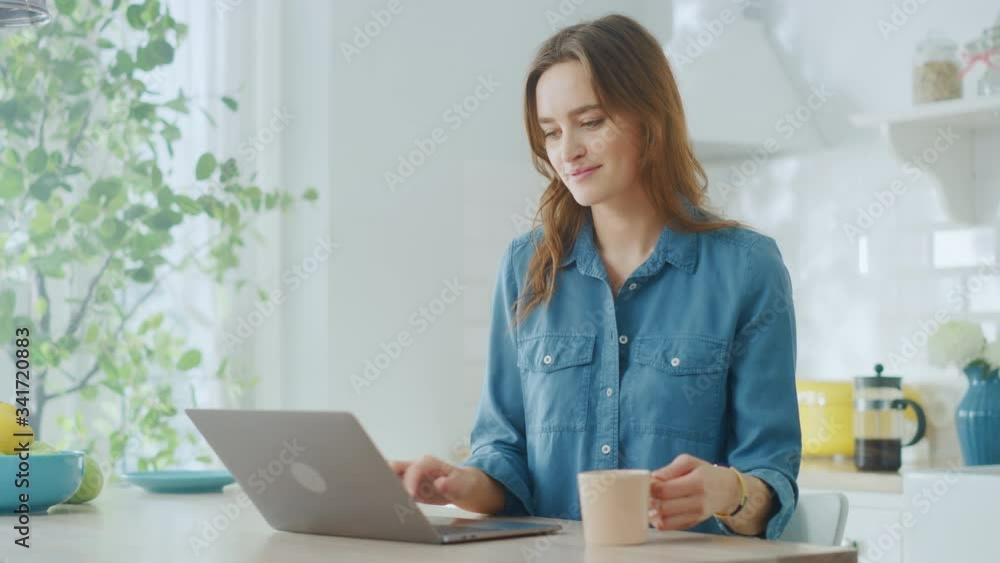 Young Attractive Woman Using Laptop Computer and Drinking Morning Cup of Coffee or Tea While Wearing a Jeans Shirt. Brunette Female Sitting in a Modern Cozy Kitchen. Freelancer Working from Home. Young Attractive Woman Using Laptop Computer and Drinking Morning Cup of Coffee or Tea While Wearing a Jeans Shirt. Brunette Female Sitting in a Modern Cozy Kitchen. Freelancer Working from Home.