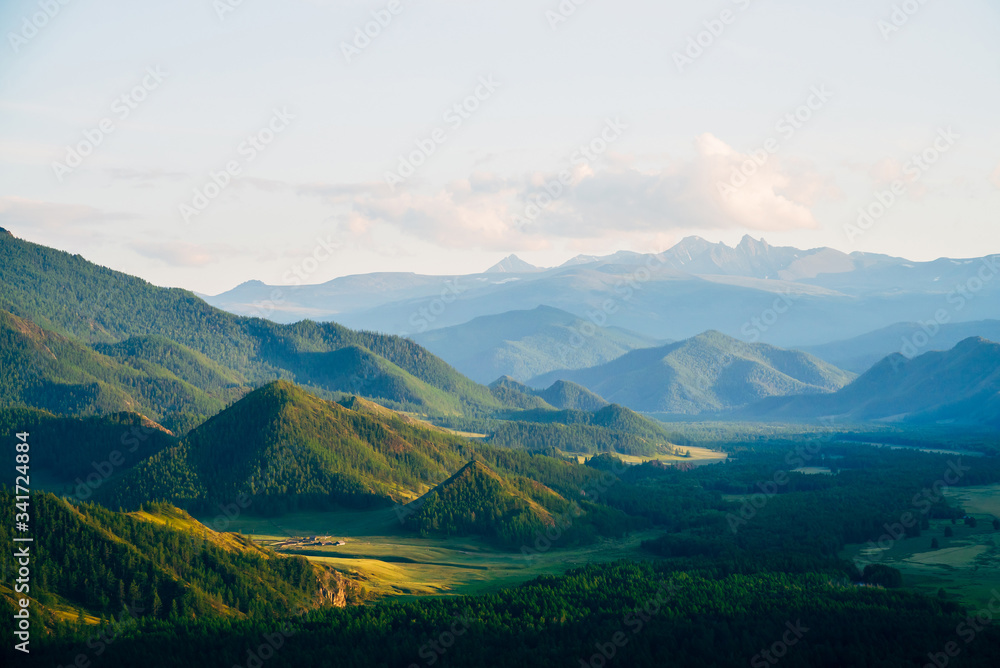 Wonderful mountain land with forest and small houses in sunlight. Scenic view to vast expanses of rolling hills and great mountain ridge on horizon. Immense distances of wavy relief in evening light.