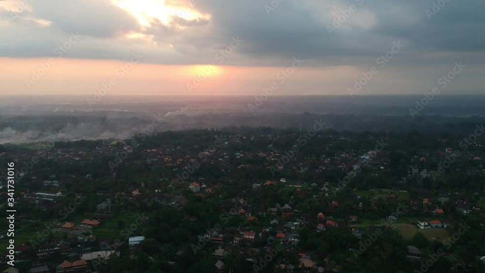 the quadrocopter takes off over Ubud in Bali, the artists road and red roofs of houses are visible, it is moving away from the sunset, the whole sky is in clouds and the yellow-orange sun is visible the quadrocopter takes off over Ubud in Bali, the artists road and red roofs of houses are visible, it is moving away from the sunset, the whole sky is in clouds and the yellow-orange sun is visible