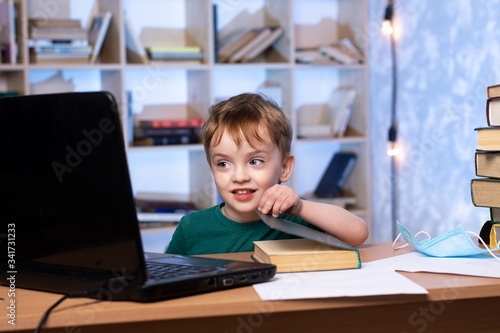 little boy sits at a desk with a laptop and opens a book. bookcase on the background. distance learning. online lessons