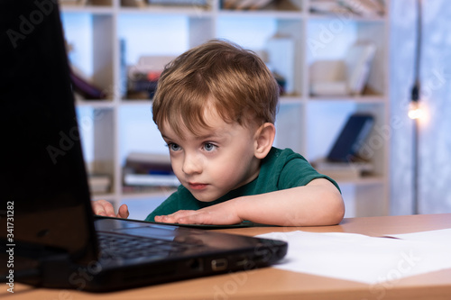 a three-year-old child is sitting at a desk and looking at a laptop. bookcase on the background. home schooling.
