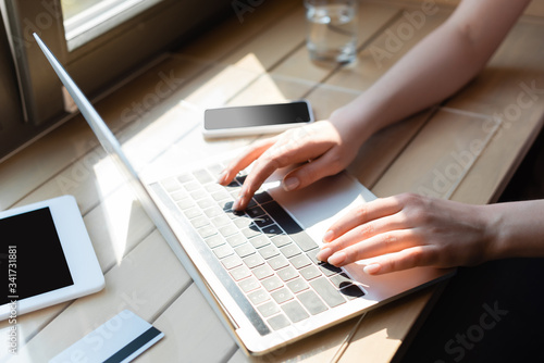 cropped view of woman typing on laptop keyboard near credit card and gadgets with blank screen
