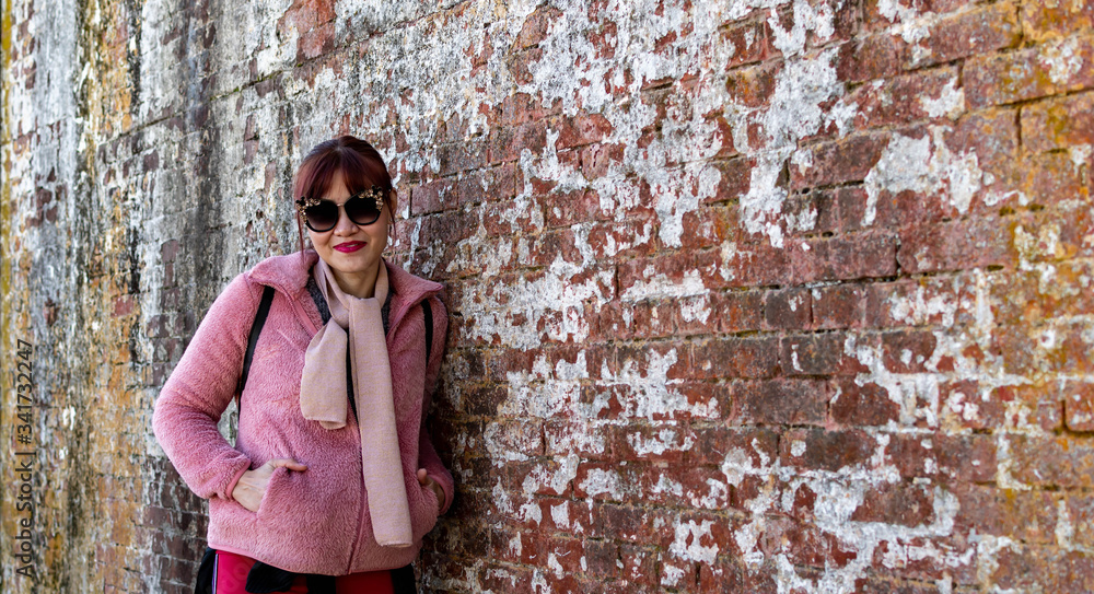 Asian female wearing jacket and eye glasses standing beside an old brick wall
