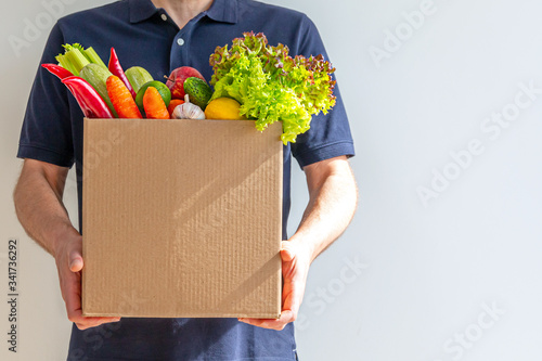 Food delivery service - man with a cardboard box of fresh vegetables on a gray background. Copy space