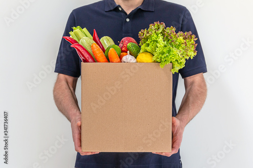 Food delivery service - man in blue uniform with a big box of fresh and raw vegetables. Order from a courier at home, express delivery, food delivery, online shopping concept
