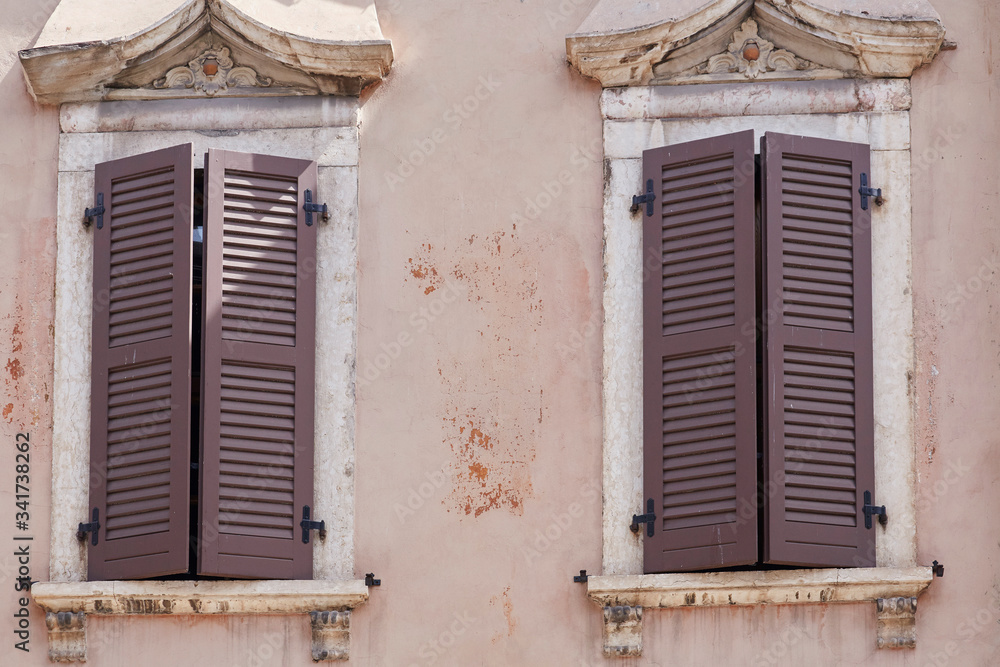 Two Italian windows on the pink flaky wall facade with closed wooden brown color classic shutters Two Italian windows on the pink flaky wall facade with closed wooden brown color classic shutters