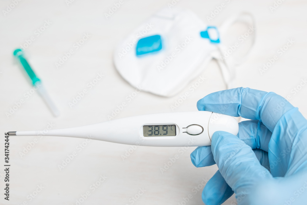 Concept: The doctor holds a thermometer showing high temperature. In the background lies an ffp3 respirator, and a syringe. Thermometer shows a temperature of 38.9 Concept: The doctor holds a thermometer showing high temperature. In the background lies an ffp3 respirator, and a syringe. Thermometer shows a temperature of 38.9