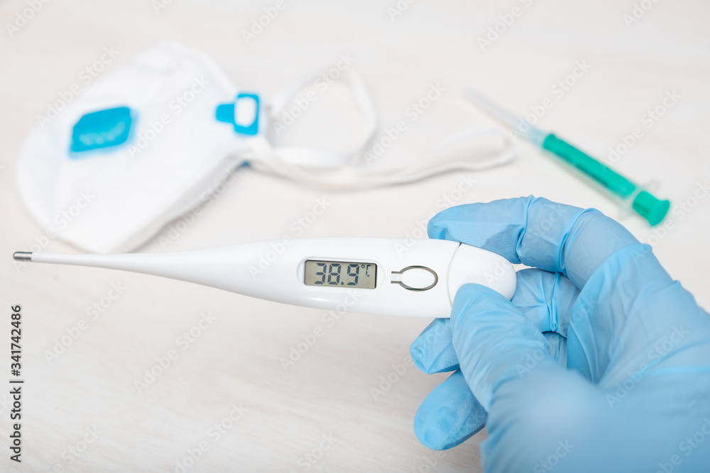 Concept: The doctor holds a thermometer showing high temperature. In the background lies an ffp3 respirator, and a syringe. Thermometer shows a temperature of 38.9 Concept: The doctor holds a thermometer showing high temperature. In the background lies an ffp3 respirator, and a syringe. Thermometer shows a temperature of 38.9