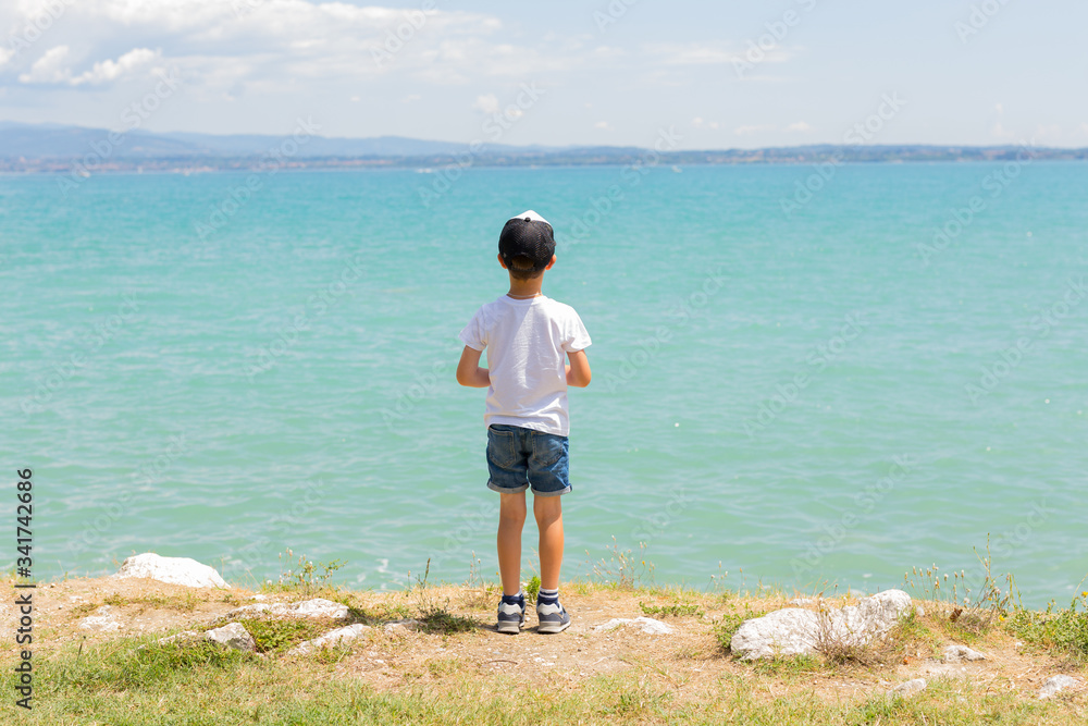 Boy stands on coast of lake, sunny daytime. Healthy leisure. Summer. Calmness. Nature. Panoramic view. Blue clear water. Landscape. Postcard. Travel. Summer family vacation. 
