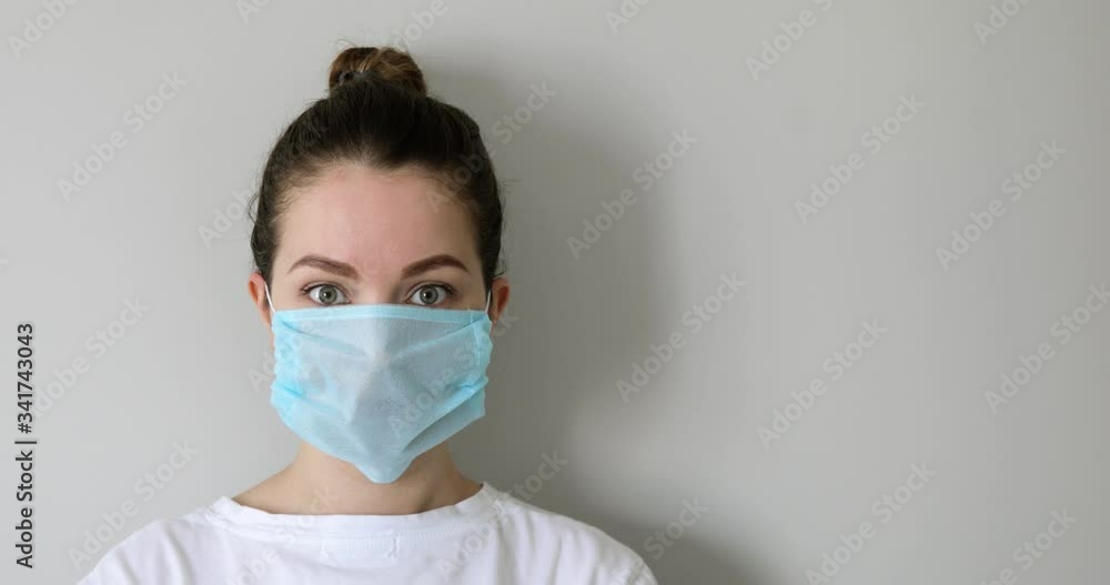 Brunette young woman putting on protective mask on face in coronavirus pandemic on grey background. Girl in protective medical mask wearing in white t-shirt. COVID-19 pandemic, epidemic, quarantine. Brunette young woman putting on protective mask on face in coronavirus pandemic on grey background. Girl in protective medical mask wearing in white t-shirt. COVID-19 pandemic, epidemic, quarantine.