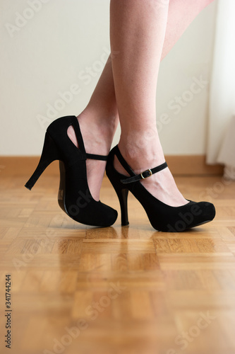 Young Caucasian woman pale white legs and black high heel shoes standing on wooden tile floor close to large window with drapes no face unrecognizable