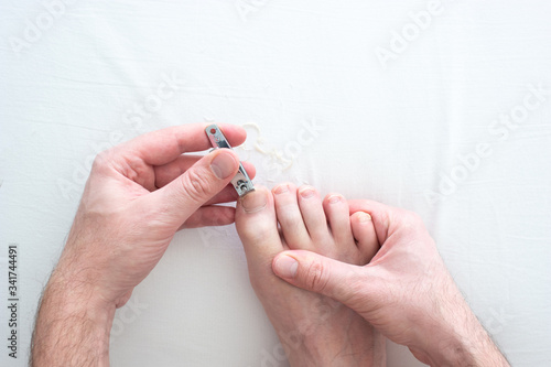 Caucasian male hand and foot cutting his toenails with a metal nail clipper on a white surface top view close up shot photo