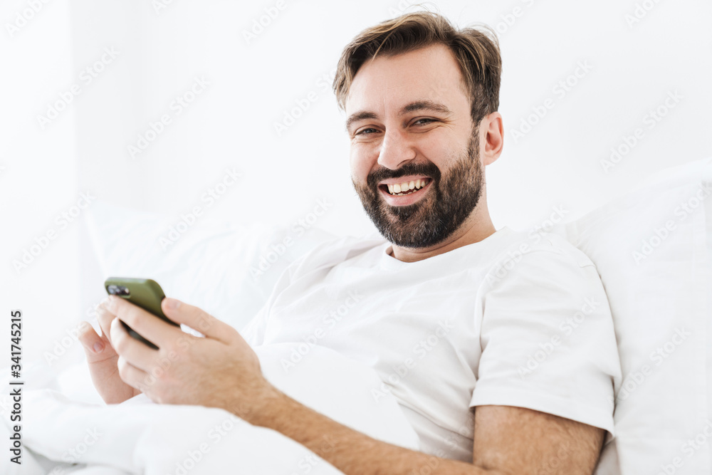 Image of young unshaven caucasian man smiling and using cellphone while lying in bed at home Image of young unshaven caucasian man smiling and using cellphone while lying in bed at home