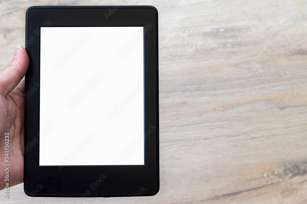 A modern black electronic book with a white blank empty screen in human hand against a blurred wooden floor background. Mockup tablet closeup with copy space A modern black electronic book with a white blank empty screen in human hand against a blurred wooden floor background. Mockup tablet closeup with copy space