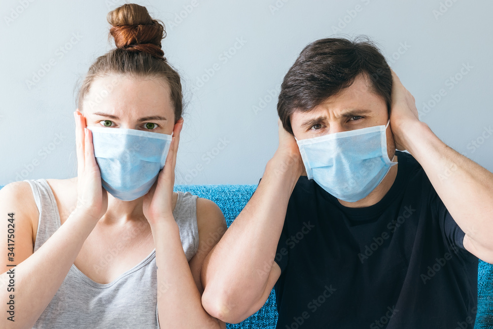 Married couple in blue medical face masks sitting on the couch, holding their heads with their hands. Man and woman in shock, family of two, stay at home during quarantine