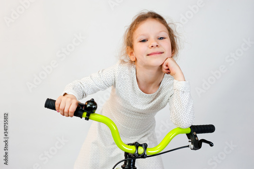 Beautiful little girl with curly hair in a light dress on a new bright bicycle. Shot on a light background.