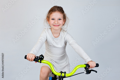 Beautiful little girl with curly hair in a light dress on a new bright bicycle. Shot on a light background.