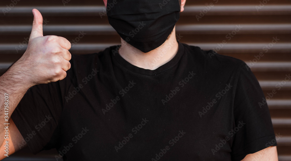 Young man in black t-shirt and mask isolated on dark wall background. Healthy lifestyle, treating sick patients, concept of cold season. They mock copy space. Wear a sterile face mask, thumbs up Young man in black t-shirt and mask isolated on dark wall background. Healthy lifestyle, treating sick patients, concept of cold season. They mock copy space. Wear a sterile face mask, thumbs up