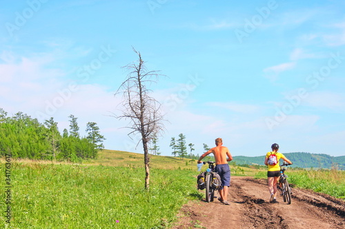 Two cyclist, man and woman, biking on a bike lane inside a park with flower field and blue sky.