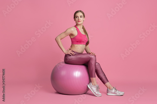 Flexible. Beautiful young female athlete practicing in studio, monochrome pink portrait. Sportive fit caucasian model training with fitball. Body building, healthy lifestyle, beauty and action concept