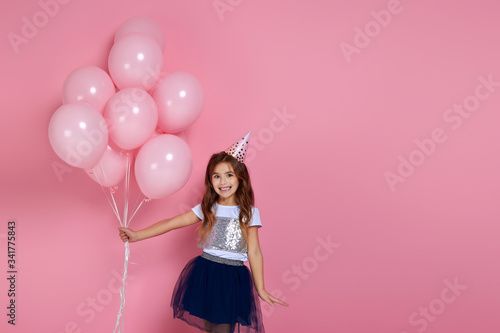 Beautiful happy little child girl in dress and birthday hat celebrating with pastel pink air balloons isolated on pink background. birthday party. copy space