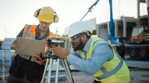 Inside of the Commercial / Industrial Building Construction Site: Engineer Surveyor Takes Measures with Theodolite, Using Digital Tablet Computer. In Background Skyscraper Formwork Frames and Crane 