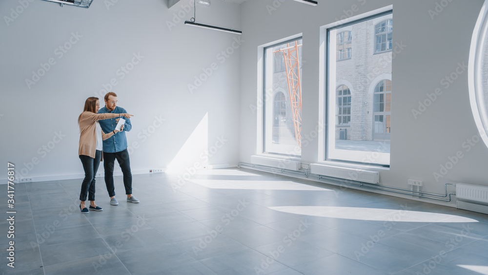Fototapeta premium Young Hipster Man and Female Stand in an Empty White Office and Map it with an Augmented Reality Software on a Tablet. Sunlight Shines Through Big Windows.