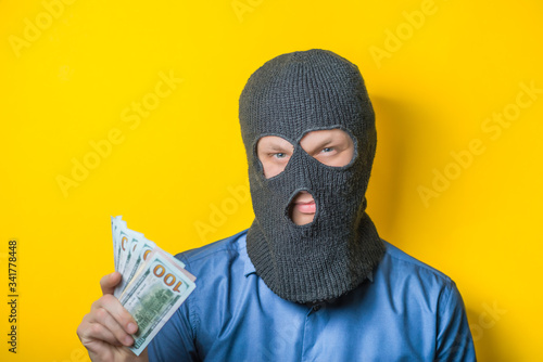 man close up thief in a mask and a blue shirt on a yellow background looks slyly to the camera. Mimicry. Gesture. photo Shoot