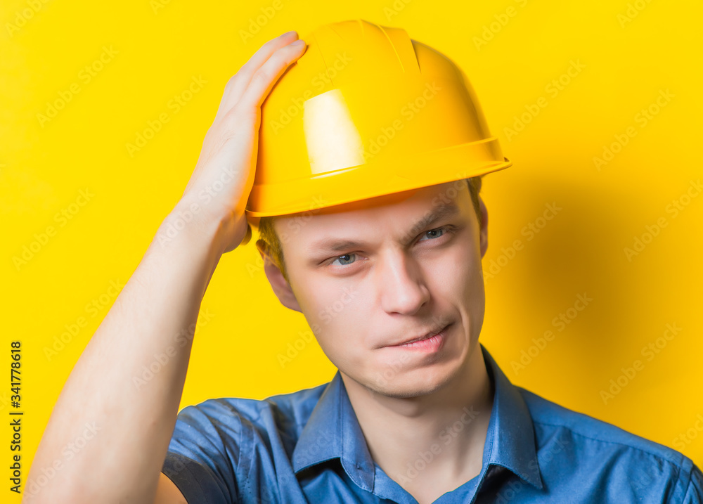 Young man close-up in yellow construction helmet and a blue shirt on a yellow background headache, holding his head by hand. Mimicry. Gesture. photo Shoot Young man close-up in yellow construction helmet and a blue shirt on a yellow background headache, holding his head by hand. Mimicry. Gesture. photo Shoot