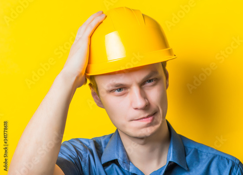 Young man close-up in yellow construction helmet and a blue shirt on a yellow background headache, holding his head by hand. Mimicry. Gesture. photo Shoot
