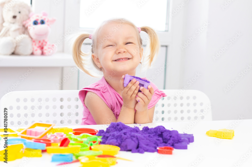 Girl plays kinetic sand in quarantine. Blond beautiful girl smiles and plays with purple sand on a white table. Girl plays kinetic sand in quarantine. Blond beautiful girl smiles and plays with purple sand on a white table.
