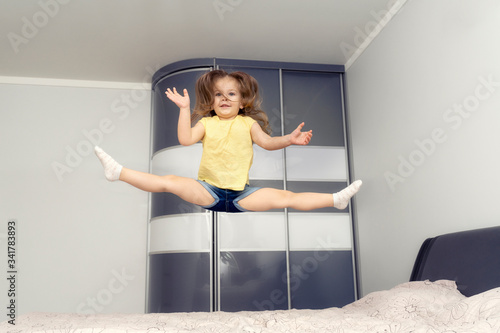 Fototapeta Naklejka Na Ścianę i Meble -  a little girl of 4 years old in a yellow T-shirt bounces on the bed and does the twine. quarantined little gymnast