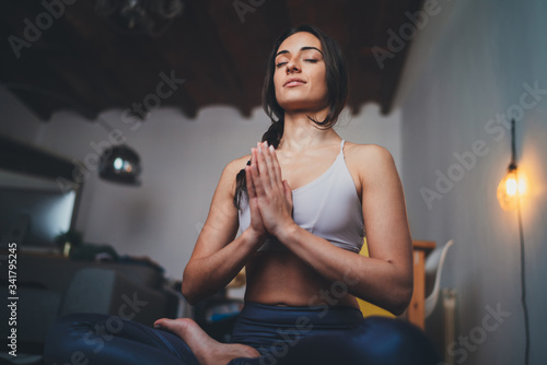 Young healthy beautiful woman in sportive top and leggings practicing yoga at home sitting in lotus pose on yoga mat meditating smiling relaxed with closed eyes, Mindfulness meditation concept