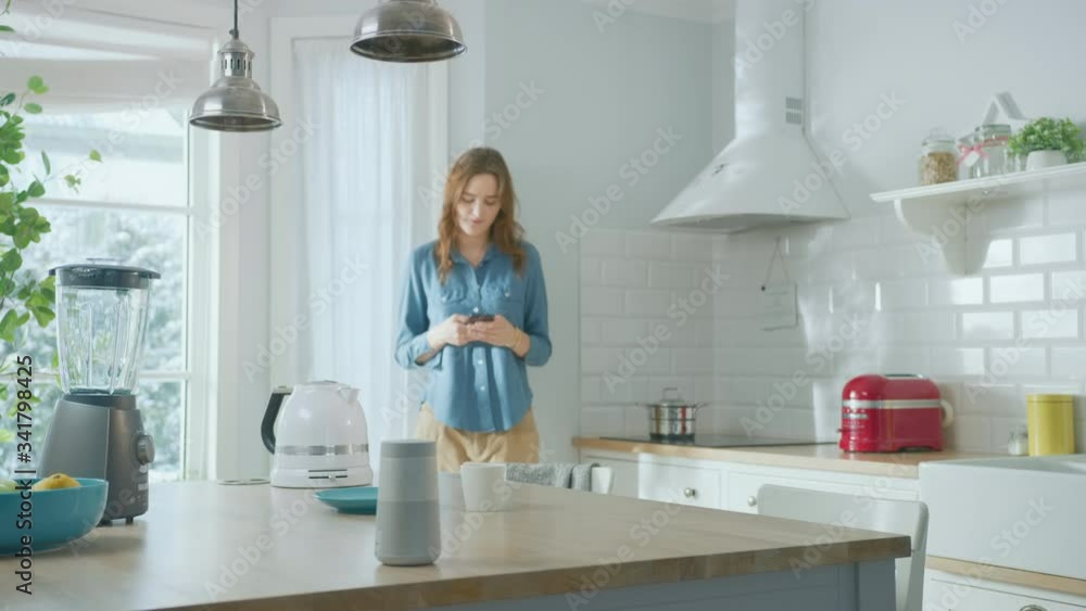 Beautiful Female Using Smartphone in Bright Sunny Kitchen. She is Connecting Electrical Appliances with Her Smart Home Application Software. Attractive Girl Browsing Internet, Social Networking Apps. Beautiful Female Using Smartphone in Bright Sunny Kitchen. She is Connecting Electrical Appliances with Her Smart Home Application Software. Attractive Girl Browsing Internet, Social Networking Apps.