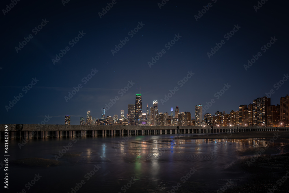 Building lights reflecting off of standing water adjacent to corrugated steel sheet piling and concrete pier with the Chicago Skyline on the horizon and stars and planes in the sky above.
