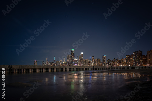 Building lights reflecting off of standing water adjacent to corrugated steel sheet piling and concrete pier with the Chicago Skyline on the horizon and stars and planes in the sky above.