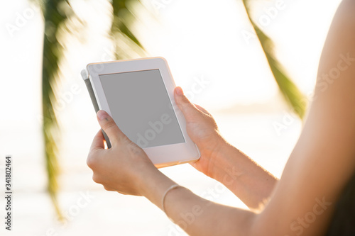 Woman holding e-reader outdoors by the sea with a palm trees on background. Relaxing and enjoying with a reading a favorite books outdoor in a travel.