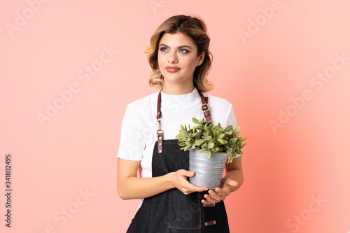 Russian gardener girl holding a plant isolated on pink background standing and looking to the side