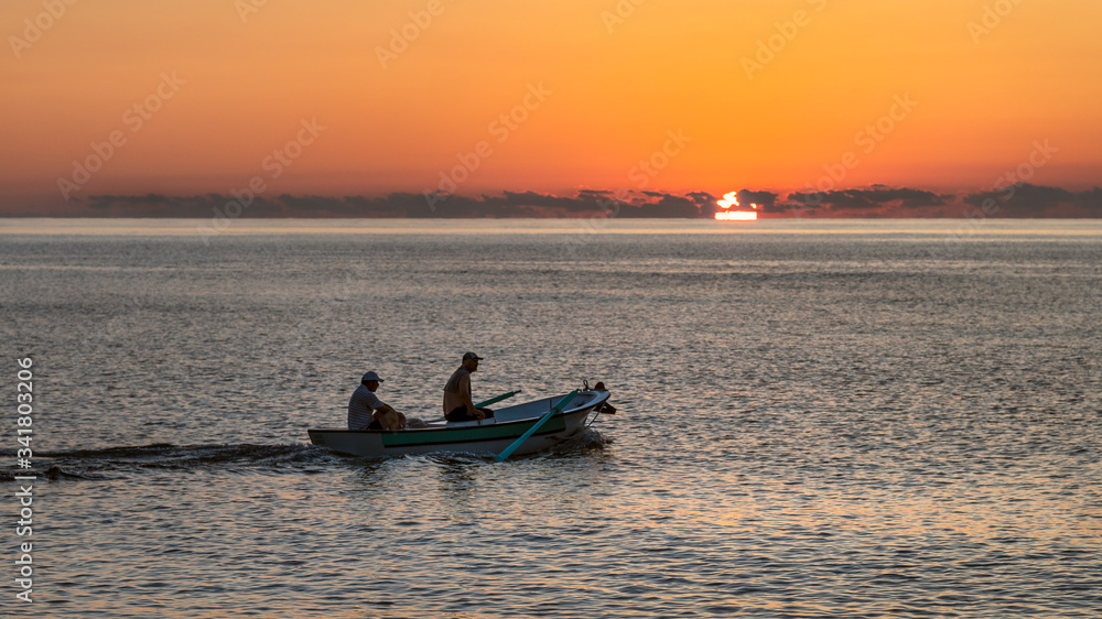 Two fishermen passing by the boat with the sun set at sunset on the horizon. They're returning home to their prey.