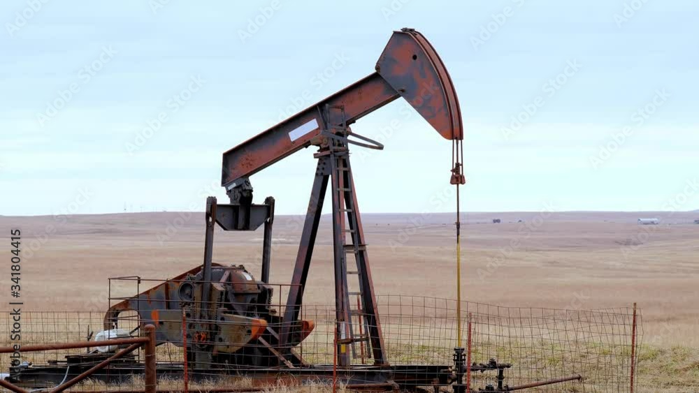 Oil Well Pump Jack pumping crude oil for fossil fuel energy. American Petroleum Oil and Gas Industry equipment extracting from a field on a prairie in the United States of America. Zoom out.