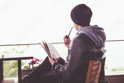 Back view on young black haired man witn piercing reeding a book and smoking tabacco pipe in outdoor cafe